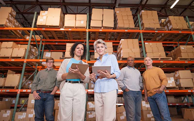 Five workers stand in a warehouse in front of shelves filled with boxes, holding clipboards and discussing inventory, related to IC DISC activities.
